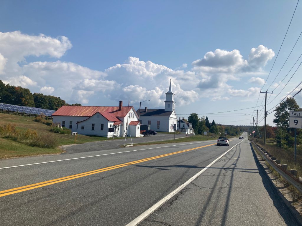 Looking along road, there are some buildings on the left side, including a church, and a white building with a shiny copper roof.  