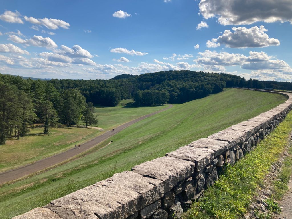 Grassy slop of a large earthen dam, with a short stone wall running along the top of it.  Some people can be seen at a distance, walking on a path at the bottom of the slope.