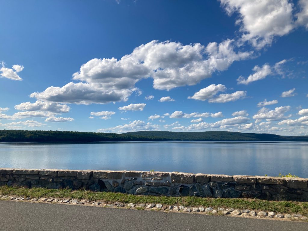 Water of Quabbin reservoir, with top surface of dam in foreground, and hills in background, clouds in sky