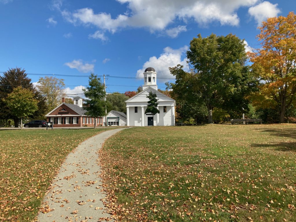 Grassy town common with paved sidewalk going through the middle.  At the far side of the grass the sidewalk leads to a white building with a cupola on top.  There is a shorter brick building next to the white one, and trees among and behind the buildings.  There are numerous dry, fallen leaves on the grass.