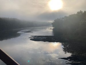 View of a river in light morning fog, with the sun still low in the sky.  Trees line each side of the water, and there is a bit of metal bridge superstructure visible in the lower left corner.