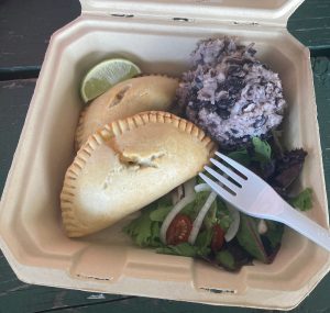 Takeout food tray containing 2 baked empanadas, a ball of black beans and rice, some salad, a lime wedge, and a plastic fork.