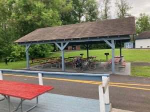 Wooden picnic shelter, with posts painted grayish blue, and several picnic tables under it, as well as my mike with luggage on the back.  There's grass and trees beyond the shelter, and in front of it is the paved trail passing by, and a picnic table near the dock.