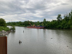 View of canal water with a barge parked on the far side, and trees lining the far bank.  There is construction equipment visible on the barge.