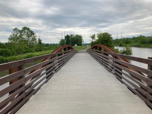 Looking along trail bridge with metal superstructure and rails, and concrete deck.  There are trees and brush to the left of the trail, and canal water to the right.