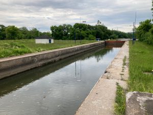 Looking along small concrete lock filled with water, with metal gate visible at far end.  There is grass on both sides of the lock, with a small building visible in the medium distance, and trees beyond.