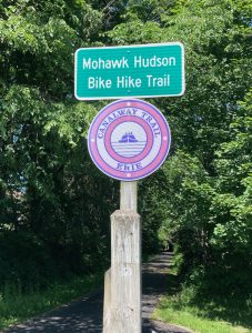 Sign post with green sign reading "Mohawk Hudson Bike Hike Trail" in white lettering, as well as pink and white sign reading "Canalway Trail Erie".  There are trees and trail pavement in the background.