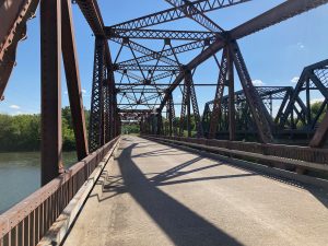 Looking along bridge pavement with rusty iron superstructure, some water and trees visible to the left side.  Another similar bridge is visible to the right.  The sun is casting shadows of the girders on the pavement.